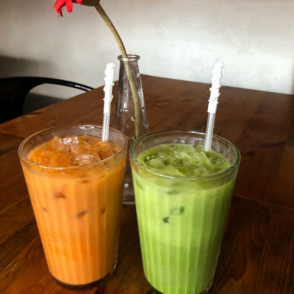 Two iced Thai drinks, one orange and one green, on a wooden table with a flower.